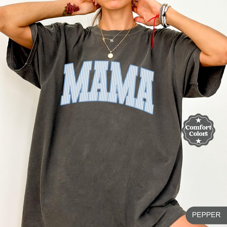 以下が含まれることがあります： 大きな「MAMA」の文字がライトブルーの縦縞でプリントされた、ダークグレーのTシャツ。Tシャツは半袖でゆったりとしたフィット感。右側にはComfort Colorsのロゴが見えます。