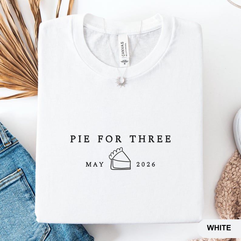 Może przedstawiać: Biała bluza z okrągłym dekoltem z napisem "PIE FOR THREE" nad "MAY 2026" i grafiką kawałka ciasta. Bluza ma małą zawieszkę w kształcie słońca i metkę "Canvas".