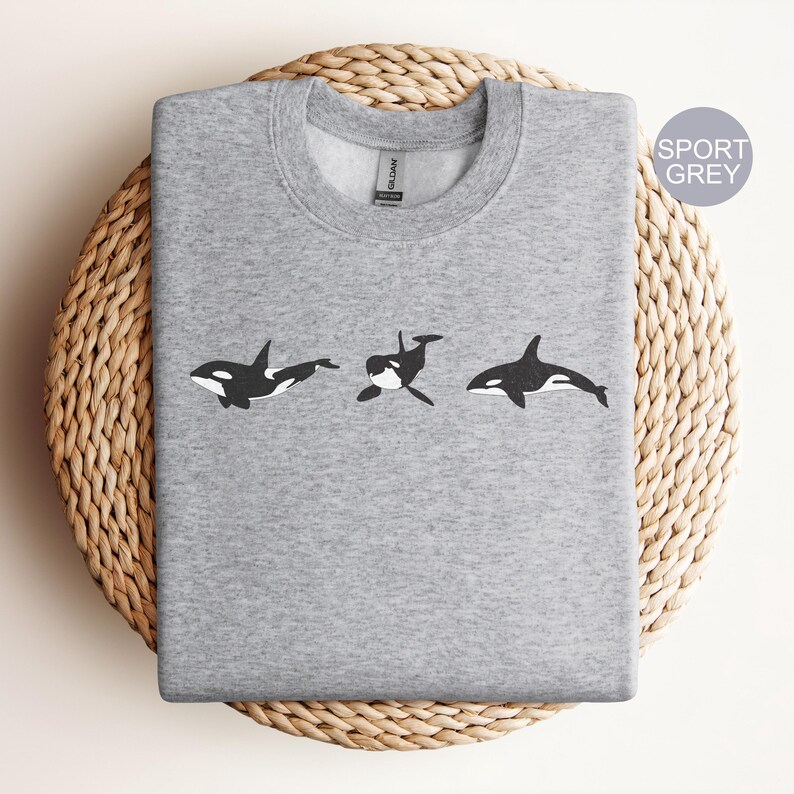 May include: A gray sweatshirt with a black and white graphic of three orcas swimming in a row. The sweatshirt is on a woven wicker surface. The text "SPORT GREY" is in the upper right corner of the image.