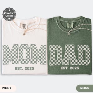 May include: Two Comfort Colors t-shirts, one ivory and one moss green, with the words "MOM" and "DAD" in a checkered pattern. Each shirt has "EST. 2025" printed below the text. The ivory shirt has a silver necklace.