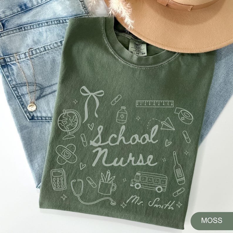 May include: Moss green t-shirt with the words "School Nurse" and related graphics, including a globe, bandage, and school bus. The name "Mr. Smith" is printed below the design. The shirt is laid on top of blue jeans and a tan hat.