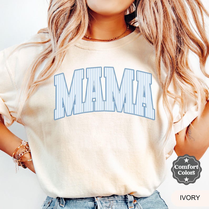 以下が含まれることがあります： アイボリー色のスウェットシャツ。ライトブルーの縦縞模様の文字で「MAMA」と書かれています。スウェットシャツはComfort Colorsブランドです。「IVORY」の文字がComfort Colorsのロゴの下に印刷されています。