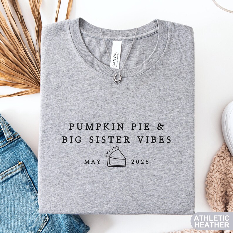 Może przedstawiać: Szara koszulka typu athletic heather z napisem "PUMPKIN PIE & BIG SISTER VIBES" i datą "MAY 2026". Widoczny naszyjnik z zawieszką w kształcie słońca. Koszulka leży obok pary dżins&oacute;w.