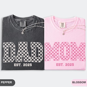 May include: Two t-shirts, one pepper-colored and one blossom-colored, each with a necklace. The pepper shirt says "DAD" in a checkered pattern, and the blossom shirt says "MOM" in a checkered pattern. Both shirts say "EST. 2025".