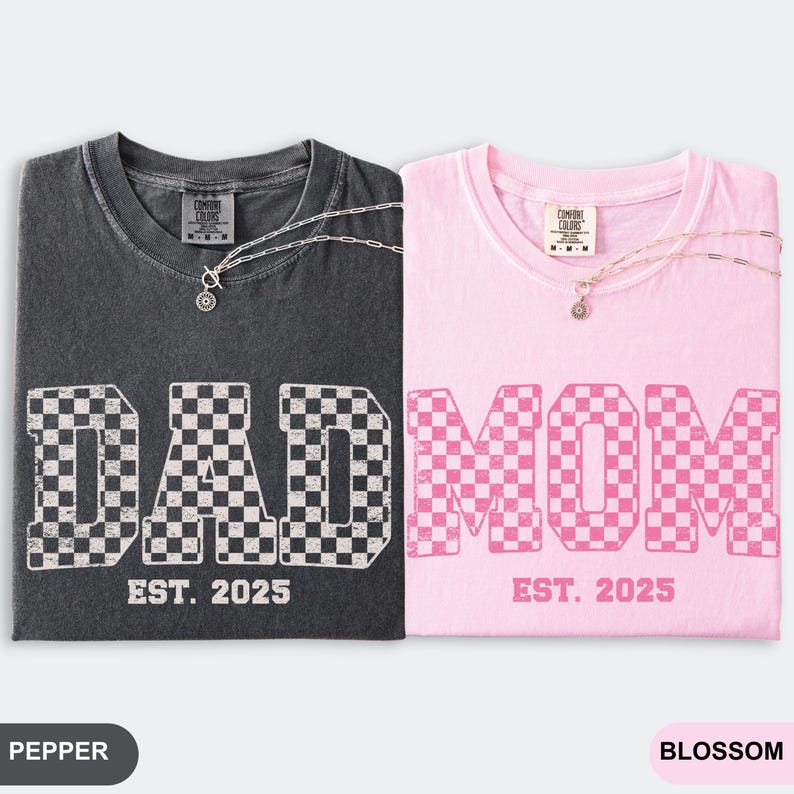 May include: Two t-shirts, one pepper-colored and one blossom-colored, each with a necklace. The pepper shirt says "DAD" in a checkered pattern, and the blossom shirt says "MOM" in a checkered pattern. Both shirts say "EST. 2025".