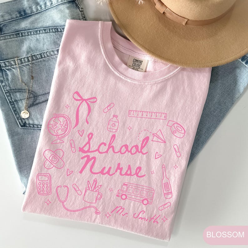 May include: A light pink t-shirt with a pink graphic design featuring school-related icons and the words "School Nurse." The design includes a bow, globe, band-aid, calculator, stethoscope, bus, and other elements. The shirt is folded and placed on top of blue jeans and a tan hat.