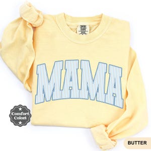 Könnte beinhalten: Ein hellgelbes Langarm-Sweatshirt mit Rundhalsausschnitt und dem Wort MAMA in einer großen, geschwungenen Schriftart im College-Stil. Die Buchstaben sind mit einem hellblau-weißen vertikalen Nadelstreifenmuster gefüllt und von einem dünnen blauen Rand umgeben.