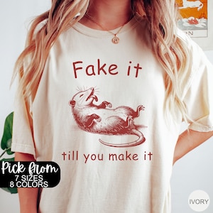 May include: A cream-colored t-shirt with a red graphic of a possum lying on its back with the text "Fake it till you make it".