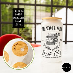 K&ouml;nnte beinhalten: Ein gefrosteter Glasbecher mit Bambusdeckel und Strohhalm. Der Becher hat ein schwarz-wei&szlig;es Design mit dem Text "The Novel Nook Book Club" und einem Stapel B&uuml;cher. Der Text "Lost in stories, found in friendship" befindet sich unter dem B&uuml;cherstapel. Der Becher steht auf einer wei&szlig;en Oberfl&auml;che.