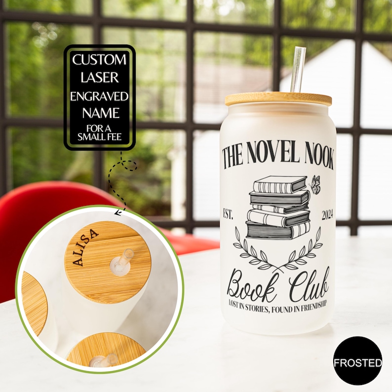 K&ouml;nnte beinhalten: Ein gefrosteter Glasbecher mit Bambusdeckel und Strohhalm. Der Becher hat ein schwarz-wei&szlig;es Design mit dem Text "The Novel Nook Book Club" und einem Stapel B&uuml;cher. Der Text "Lost in stories, found in friendship" befindet sich unter dem B&uuml;cherstapel. Der Becher steht auf einer wei&szlig;en Oberfl&auml;che.