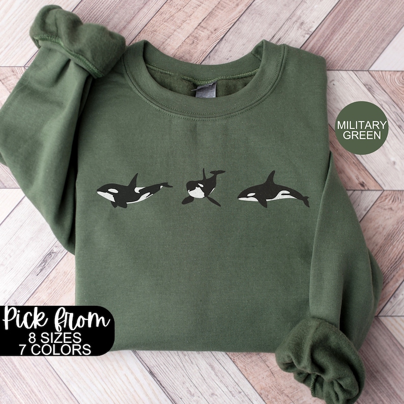 May include: A military green sweatshirt featuring a graphic of three orcas. The text "Pick from 8 sizes 7 colours" and "Military Green" are printed on the sweatshirt.