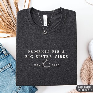 Może przedstawiać: Ciemnoszara melanżowa koszulka z białym napisem "PUMPKIN PIE & BIG SISTER VIBES MAY 2026". Widoczny naszyjnik z zawieszką w kształcie słońca. Koszulka jest złożona na białej powierzchni obok niebieskich dżins&oacute;w i beżowego swetra.