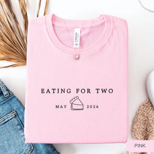 Może przedstawiać: Jasnor&oacute;żowa koszulka z czarnym napisem "EATING FOR TWO", a także "MAY 2026" i ilustracją ciasta. Koszulka jest wykonana z miękkiego materiału i jest starannie złożona.