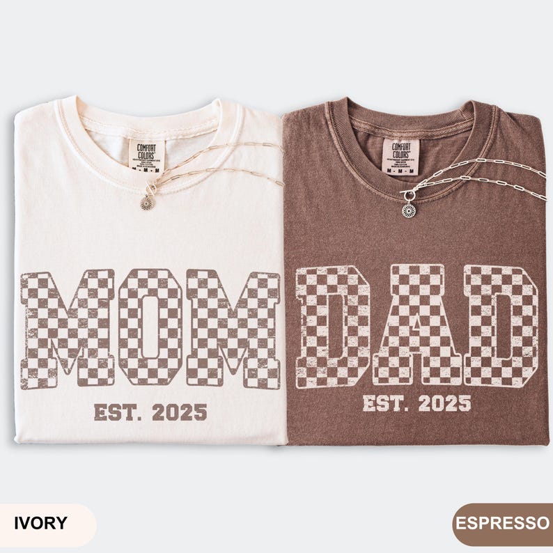 May include: Two t-shirts, one ivory and one espresso, with the words "MOM" and "DAD" in a checkered pattern. The text "EST. 2025" is below each word. Each shirt has a silver necklace.
