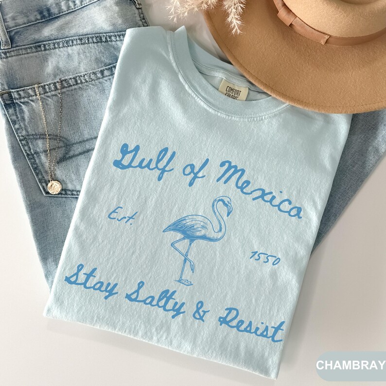 Peut inclure: T-shirt bleu clair avec un graphique de flamant rose et le texte "Gulf of Mexico Est. 1550 Stay Salty & Resist" imprim&eacute; en bleu. Le t-shirt est fait d'un mat&eacute;riau doux et confortable.