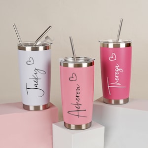 Op de afbeelding: Drie gepersonaliseerde roestvrijstalen bekers met zilveren rietjes. Eén is wit met "Jacky" in zwart script, een andere is roze met "Acheron" en de derde is magenta met "Theresa". Elk heeft een hartsymbool.