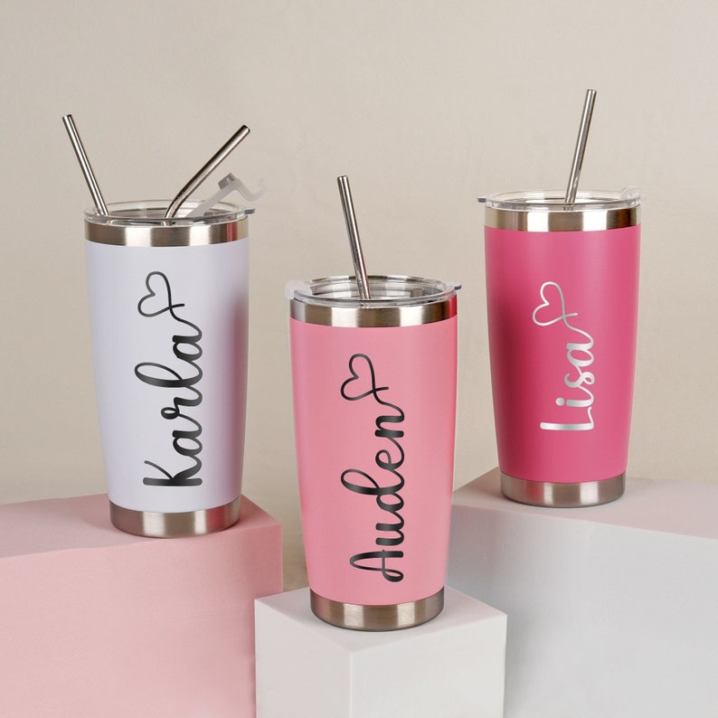 May include: Three stainless steel tumblers with personalized names, "Karla", "Auden", and "Lisa", in black script with a heart symbol. The tumblers are white, pink, and pink, and each has a stainless steel straw.
