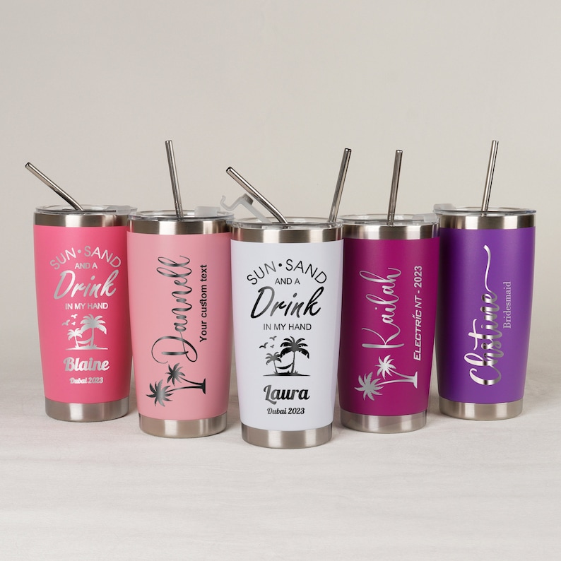 May include: Five stainless steel tumblers with lids and straws. Each tumbler is a different color: pink, light pink, white, magenta, and purple. The tumblers have text and palm tree graphics. The text includes names and phrases like "Sun, Sand, and a Drink."