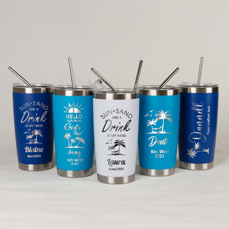 May include: Five stainless steel tumblers with various designs and text. The tumblers are blue, white, and silver. The text on the tumblers includes "Sun and a Drink", "Hello Girl's Trip", "Donti Key West 2021", "Blaine Dubai 2023", and "Laura Dubai 2023".