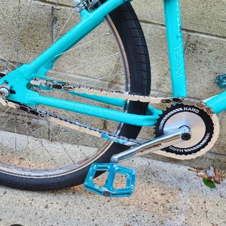Puede incluir: Primer plano de un cuadro de bicicleta turquesa con un neum&aacute;tico negro. La bicicleta presenta una cadena plateada, un engranaje negro y plateado y pedales turquesa. La palabra "TEAM" es visible en el cuadro.