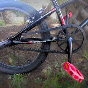May include: Black BMX bike with red accents and pink pedals. The bike features a black chain and sprocket, with the word "SHREDDER" visible on the frame. The tires are black, and the frame is made of lightweight aluminum.