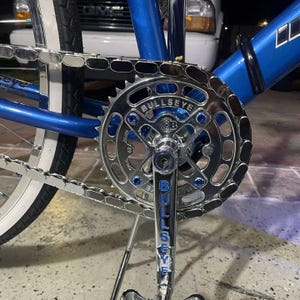Puede incluir: Primer plano de una bicicleta azul con cadena y pi&ntilde;&oacute;n plateados. El pi&ntilde;&oacute;n lleva la inscripci&oacute;n "BULLSEYE" y tiene detalles azules. El cuadro de la bicicleta es de color azul vibrante y la cadena es plateada.