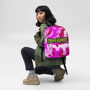 May include: Pink camouflage backpack with the text "INSANE CHAINS" in a black and yellow font. The backpack has a front pocket and black straps. The bag is made of a durable material.