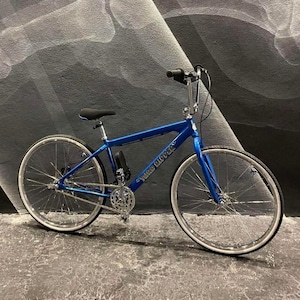 Puede incluir: Una bicicleta azul con componentes plateados y neum&aacute;ticos de flancos blancos. El cuadro tiene el texto "Tuko Ripper" impreso. La bicicleta est&aacute; sobre un fondo abstracto gris y blanco.
