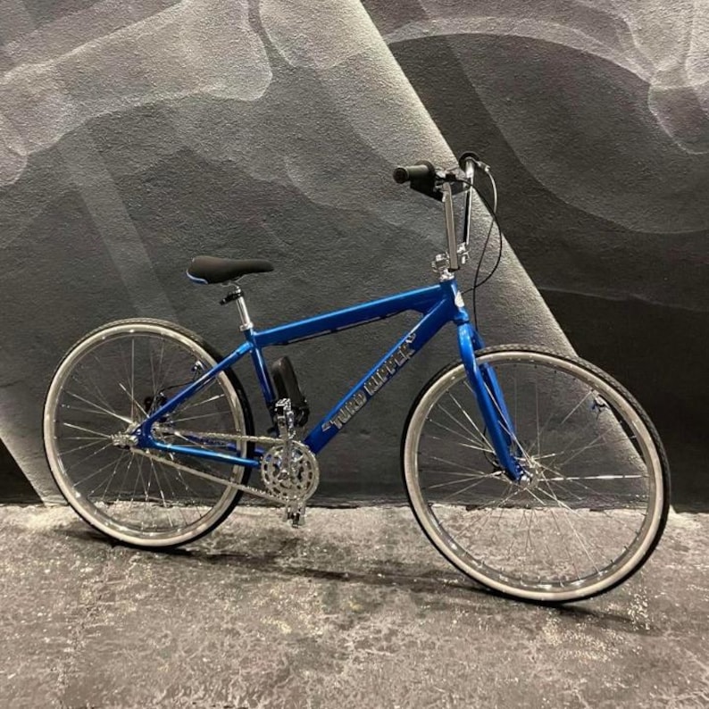 Puede incluir: Una bicicleta azul con componentes plateados y neum&aacute;ticos de flancos blancos. El cuadro tiene el texto "Tuko Ripper" impreso. La bicicleta est&aacute; sobre un fondo abstracto gris y blanco.