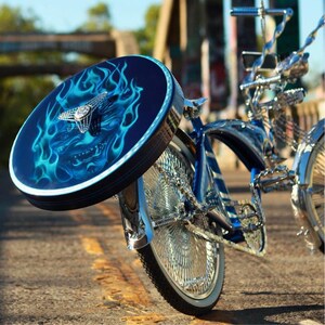 Puede incluir: Una bicicleta lowrider personalizada con una cubierta de rueda azul oscuro con un dise&ntilde;o de llamas en tonos azules y un emblema plateado. La bicicleta tiene detalles cromados y un cuadro azul, estacionada en una carretera pavimentada.