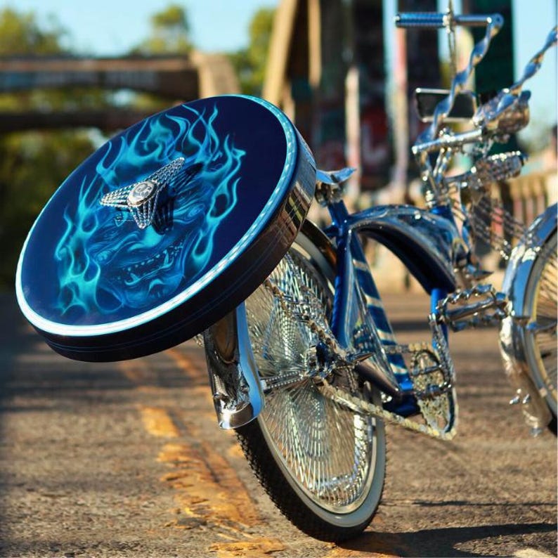Puede incluir: Una bicicleta lowrider personalizada con una cubierta de rueda azul oscuro con un dise&ntilde;o de llamas en tonos azules y un emblema plateado. La bicicleta tiene detalles cromados y un cuadro azul, estacionada en una carretera pavimentada.