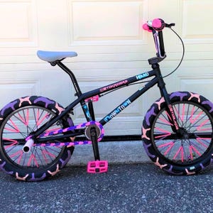 May include: A black BMX bike with pink and purple accents. The bike features pink spokes, purple and pink tires, and a light blue seat. The handlebars and pedals are pink, and the frame has the word "Downtown" on it.