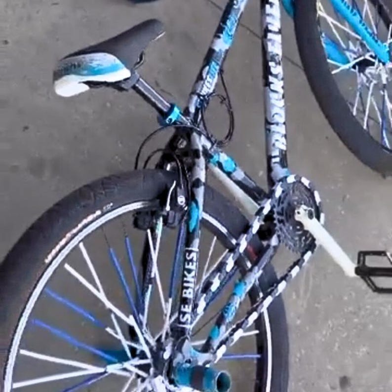 May include: A black and blue SE Bikes bicycle with white and blue accents. The bike features a black seat, blue and white rims, and a camouflage pattern on the frame. The tires are black.