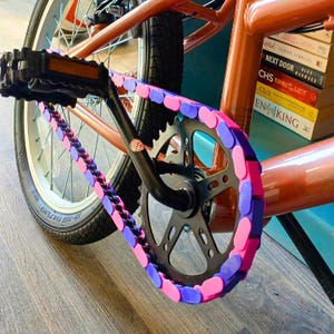 May include: Close-up of a bicycle's chain and gears. The chain is decorated with pink and purple plastic links. The bike frame is copper-colored, and the tires are black. The pedals are black, and the bike is on a wooden surface.