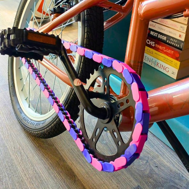 May include: Close-up of a bicycle's chain and gears. The chain is decorated with pink and purple plastic links. The bike frame is copper-colored, and the tires are black. The pedals are black, and the bike is on a wooden surface.