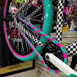 May include: Close-up of a BMX bike wheel with a teal tire and pink rim. The chain guard is black and white checkered, and the chain is teal and pink. The bike frame is white.