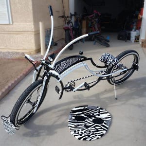 May include: A custom bicycle with a white frame, black tires, and zebra print accents on the seat, fenders, and footrests. A matching zebra print rug is in front of the bike. The bike has a unique curved design.