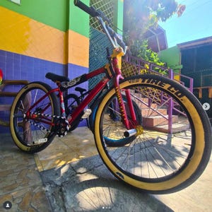 May include: A red and gold BMX bike with cream-colored tires. The bike has a black seat and handlebars. The front tire has the text "VEE TIRE CO." in white. The bike is parked outside.