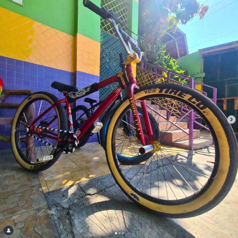 May include: A red and gold BMX bike with cream-colored tires. The bike has a black seat and handlebars. The front tire has the text "VEE TIRE CO." in white. The bike is parked outside.
