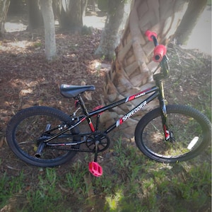 May include: A black and red HARO Shredder BMX bike with pink accents. The bike has black tires, a black seat, and pink handlebar grips and pedals. The bike is positioned on grass near a tree.