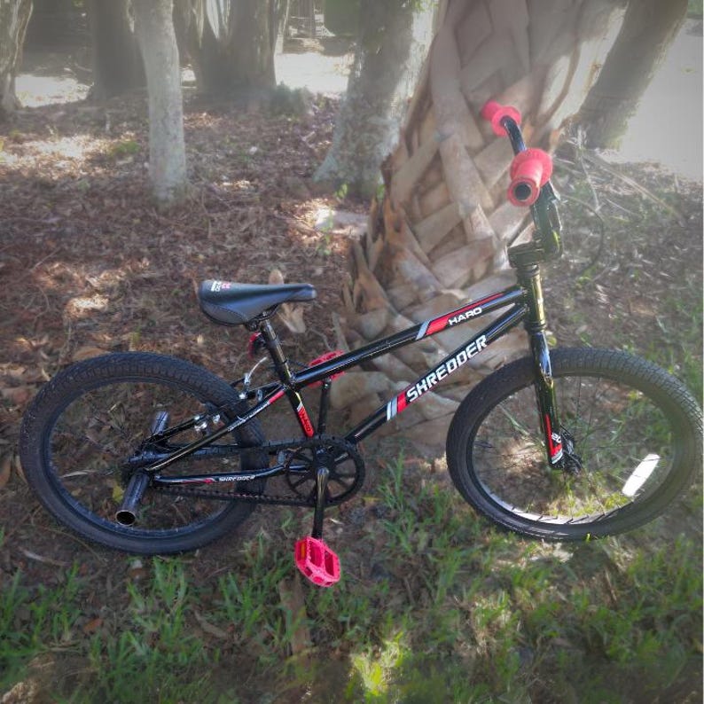 May include: A black and red HARO Shredder BMX bike with pink accents. The bike has black tires, a black seat, and pink handlebar grips and pedals. The bike is positioned on grass near a tree.
