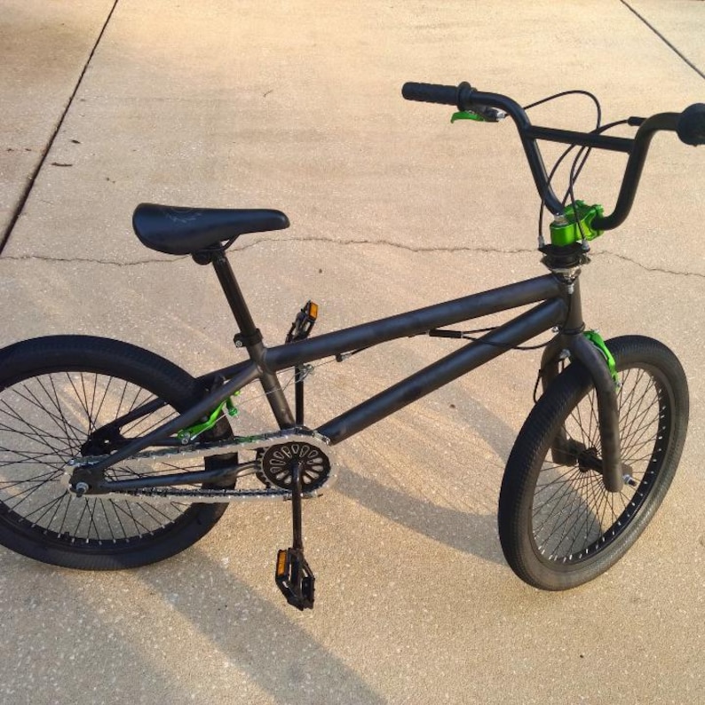 Puede incluir: Una bicicleta BMX negra con detalles verdes en el manillar, la horquilla y la cadena. La bicicleta tiene neum&aacute;ticos negros, un asiento negro y un cuadro negro. La bicicleta est&aacute; dise&ntilde;ada para acrobacias y trucos.