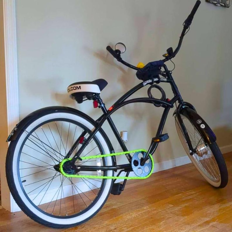 May include: Black cruiser bicycle with white-walled tires and a neon green chain. The bike has a curved frame, high handlebars, and a white seat with the word "VOLCOM" printed on it. The bike is leaning against a wall.
