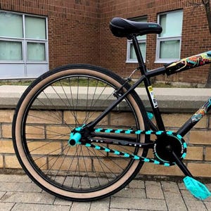 Può includere: Bicicletta nera con pneumatici color cuoio e dettagli turchese sul paracatena, sui pedali e sui mozzi delle ruote. La bici ha sella e telaio neri, con grafiche colorate sul telaio. La bici è parcheggiata su una superficie di mattoni.