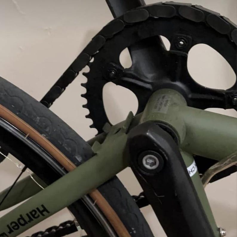 May include: Close-up of a bicycle's drivetrain, featuring a black chain and chainring. The bike frame is olive green, with a black crank arm and a portion of the tire visible. The tire has a brown sidewall and the word "Harper" is visible.