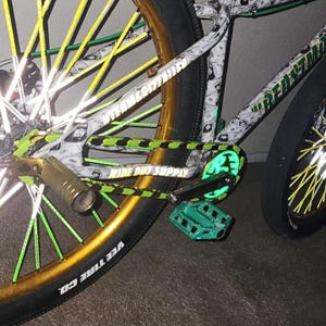Puede incluir: Primer plano de una bicicleta personalizada con llantas doradas y detalles en verde y amarillo. El cuadro est&aacute; decorado con un patr&oacute;n de billetes de d&oacute;lar. Los pedales y el protector de cadena son de color verde brillante.