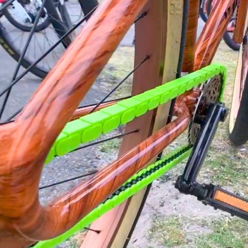 May include: Close-up of a bicycle frame with a wood grain finish. The frame has a bright green chain guard. The bicycle has black pedals and a tan tire. The bicycle is outdoors.
