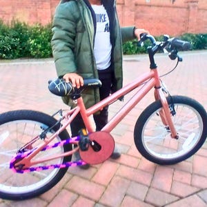 May include: A coral-colored BMX bike with black tires and white spokes. The bike features a black seat, handlebars, and pedals. The chain guard and chain are decorated with purple and pink accents. The bike is designed for recreational use.