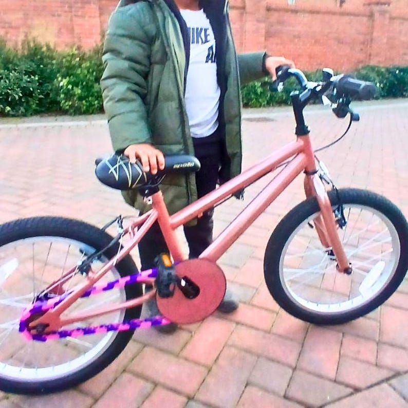 May include: A coral-colored BMX bike with black tires and white spokes. The bike features a black seat, handlebars, and pedals. The chain guard and chain are decorated with purple and pink accents. The bike is designed for recreational use.