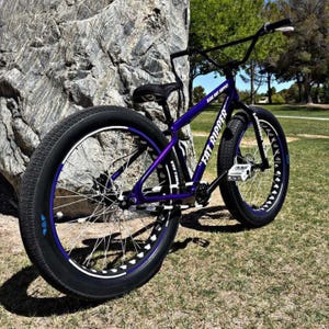May include: A purple and black "Fat Ripper" bicycle with thick tires is parked next to a large rock. The bike has a black seat, handlebars, and frame accents. The wheels have a blue and white design.